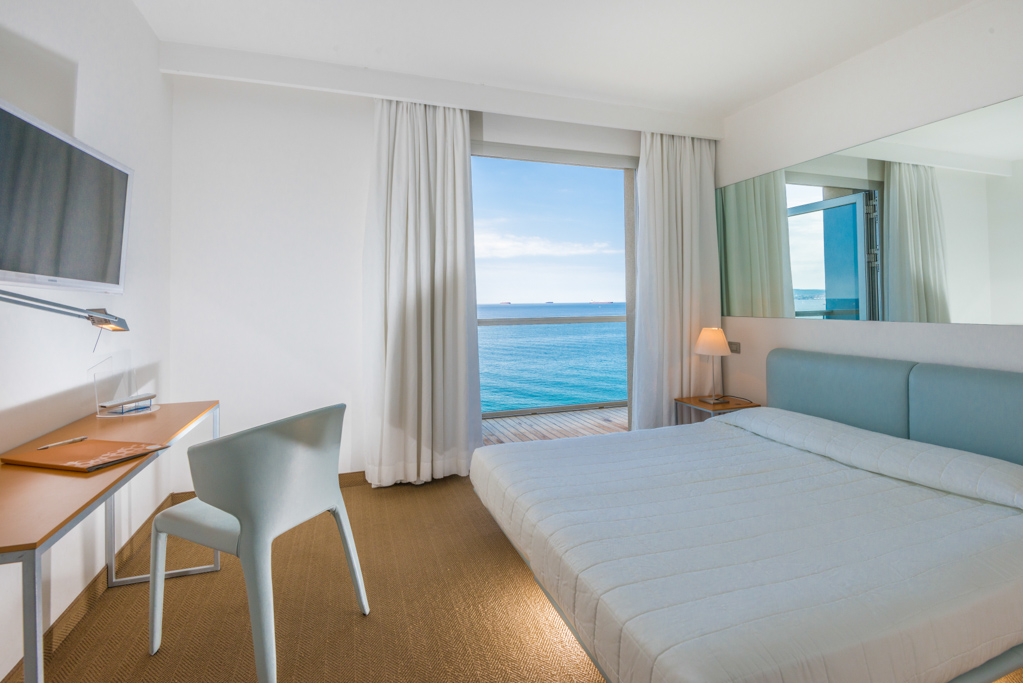 Camera d'albergo luminosa con vista sul mare attraverso una grande finestra.