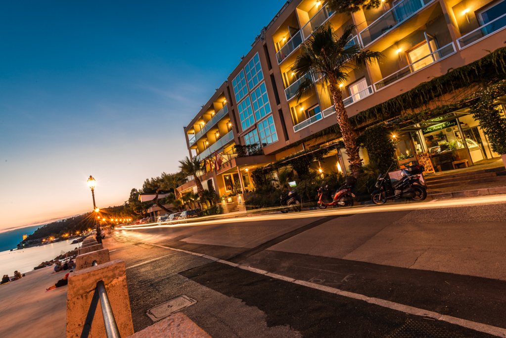 Strada costiera con hotel illuminato al tramonto e scie luminose da fari di veicoli.