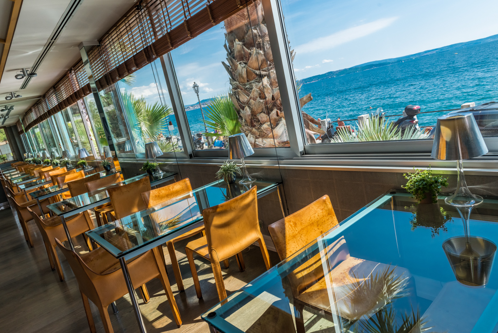 Interno di un ristorante con pareti di vetro che offre vista sul mare azzurro.